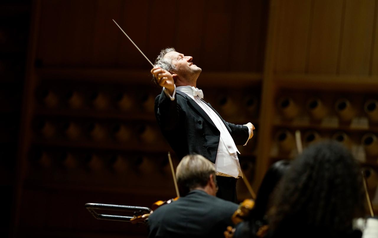 Dirigent Markus Poschner leitet mit ausdrucksvoller Geste ein Orchester während eines Konzerts im modernen Konzertsaal.