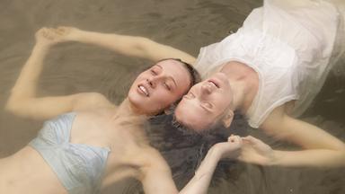 Zwei Mädchen schwimmen auf dem Rücken Kopf an Kopf in einem See und halten sich neben den Köpfen an den Händen. Das Mädchen vorne trägt blaue Uterwäsche und das andere Mädchen ein weißes Nachthemd.