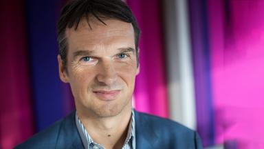 Ein Mann mit kurzen, braunen Haaren steht sich vor einem lila-farbenen Hintergrund und lächelt in die Kamera. 