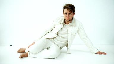 Vor weißem Hintergrund sitzt ein Mann mit weißer Kleidung und braunen Haaren auf weißem Boden.