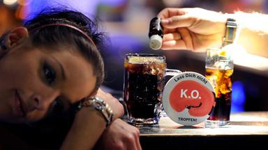 Eine Frau hat den Kopf auf den Tresen gelegt, während neben ihr eine Hand ein kleines braunes Fläschchen über ihr Glas hält.