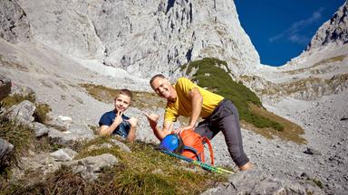 Das Bild zeigt zwei Personen in einer alpinen Landschaft. Im Vordergrund sitzen ein älterer Mann und ein Junge auf einem steinigen Untergrund, umgeben von Gräsern und niedrigen Pflanzen. Der Mann trägt ein gelbes T-Shirt und graue Hose, während der Junge ein blaues T-Shirt trägt. Beide posieren freundlich, der Junge zeigt mit beiden Händen ein Zeichen des "Shaka", während der Mann mit einer Hand das gleiche Zeichen macht.  Im Hintergrund erhebt sich eine massive Felsformation, die aus grauem Gestein besteht und steil aufsteigt. Der Himmel ist klar und blau, und im unteren Drittel des Bildes sind einige flachere, grüne Bereiche sichtbar, die mit Bäumen bewachsen sind. Das Gesamtbild vermittelt einen Eindruck von Weite und der Schönheit der alpinen Natur, während es gleichzeitig eine Elemente des Bergsteigens und der Outdoor-Aktivitäten zeigt.