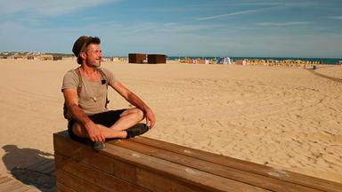 Christopher Crell im Schneidersitzt auf einer Holz-Mauer am Strand