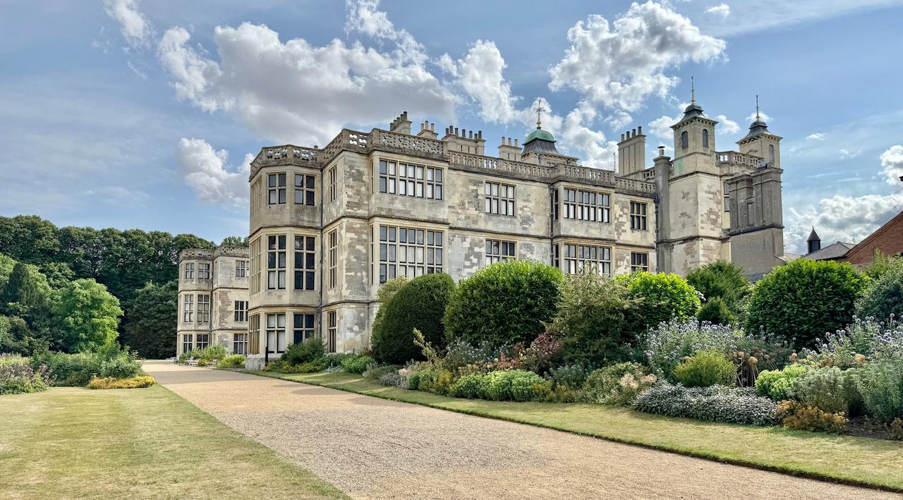 Das Bild zeigt die Ostseite des Audley End House, einem historischen Herrenhaus in England. Im Vordergrund befindet sich ein gepflegter Garten mit verschiedenen Pflanzen und Sträuchern, die in unterschiedlichen Grüntönen gehalten sind. Der Garten ist von einem schotterigen Weg umgeben, der nach links in eine größere Grünfläche übergeht.  Das Gebäude selbst hat eine eindrucksvolle Fassade mit vielen großen Fenstern, die von einer hellen Steinstruktur umgeben sind. Einige Bereiche der Wand scheinen unregelmäßig bearbeitet zu sein, möglicherweise aufgrund von Alterung oder Restaurierung. Auf dem Dach des Hauses sind Türmchen mit kleinen Spitzdächern zu sehen, die den oberen Teil des Gebäudes zieren. Der Himmel ist teilweise bewölkt und gibt dem Bild eine helle, freundliche Atmosphäre.