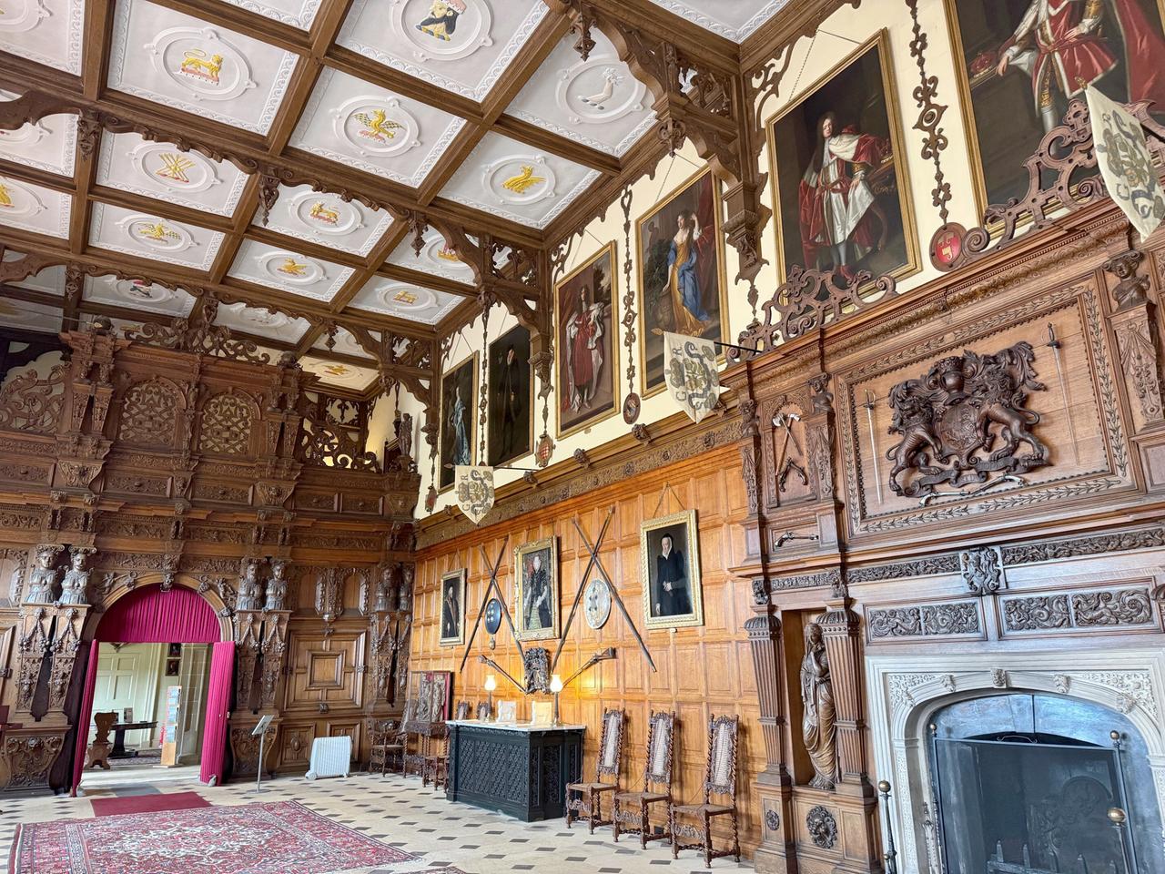 Das Bild zeigt die Great Hall im Audley End House, einem historischen Gebäude in den königlichen Gärten. Der Raum ist groß und beeindruckend, mit einer hohen Decke, die ein Muster aus weißen und holzverzierten Feldern aufweist. An den Wänden hängen mehrere Gemälde, die Personen in historischer Kleidung darstellen.   Der Raum ist mit dunklem Holz verkleidet, das kunstvoll geschnitzt ist, und es gibt große Fenster, die natürliches Licht hineinlassen. In der Mitte des Raumes liegt ein Teppich, der den Boden bedeckt, und an einer Wand steht ein großer Kamin aus Stein. Eine altmodische Bank und Stühle mit hohen Rückenlehnen sind um einen Tisch gruppiert, auf dem einige Kerzenleuchter stehen.   Ein rotes Samtvorhangstück rahmt einen Durchgang zu einem anderen Raum ein, der im Hintergrund sichtbar ist. An den Wänden sind verschiedene Wappen und Holzverzierungen angebracht. Das Gesamtbild vermittelt eine Atmosphäre von historischer Pracht und Eleganz.