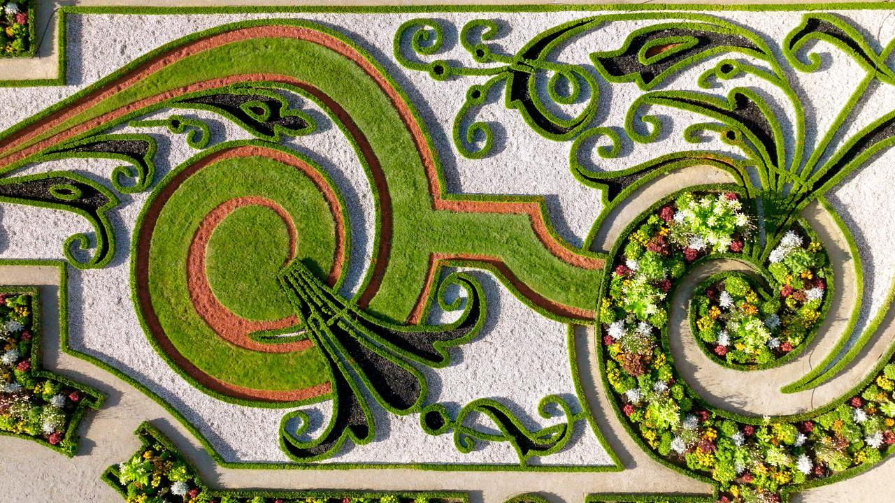 Das Bild zeigt die Blumenterrasse im Schloss Hof, die Teil der königlichen Gärten ist. Aus der Vogelperspektive betrachtet, sind die Gartenanlagen in geometrischen Mustern gestaltet. Verschiedene Grüntöne dominieren die Fläche, ergänzt durch braune und schwarze Akzente, die die Muster hervorheben.   In der Mitte sind spiralförmige und geschwungene Linien sichtbar, die mit blühenden Pflanzen in bunten Farben geschmückt sind. Diese Blumen sind in verschiedenen Gruppen angeordnet, was einen lebhaften Kontrast zu den gleichmäßig geschnittenen grünen Hecken bietet. Die gesamte Gestaltung vermittelt einen kunstvollen und gepflegten Eindruck.
