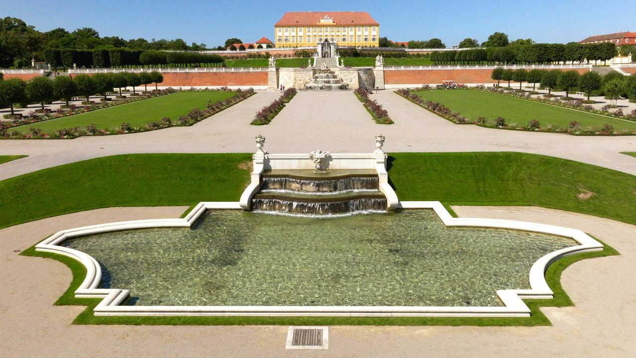 Das Bild zeigt die Kleine Kaskade im Schloss Hof, umgeben von gepflegten Gärten. Im Vordergrund befindet sich ein rechteckiger Teich mit klaren Wasser, aus dem sanft Wasser in drei Stufen in das Becken fließt. Um den Teich herum gibt es eine Einfassung aus hellem Stein. 

Im Hintergrund erhebt sich das Schloss mit einer gelben Fassade und roten Dächern, das von einer symmetrischen Gartenlandschaft mit Rasenflächen und Blumenbeeten flankiert wird. Wegezonen aus Kies führen durch die Gartenanlage, die auch mit Baumreihen versehen ist. Die gesamte Szene wird von einem klaren blauen Himmel überzogen, was eine sonnige und einladende Atmosphäre schafft.