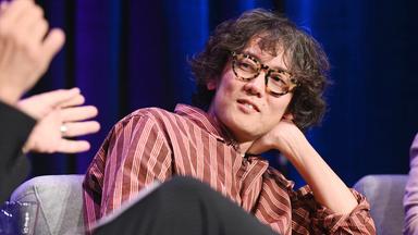 Kohei Saito - ein Japaner mit kurzen leicht graumelierten lockigen Haaren und großer Kunststoffbrille stützt sich auf eine Hand im Nacken, während er einer Person, die neben ihr sitzt, zuhört.