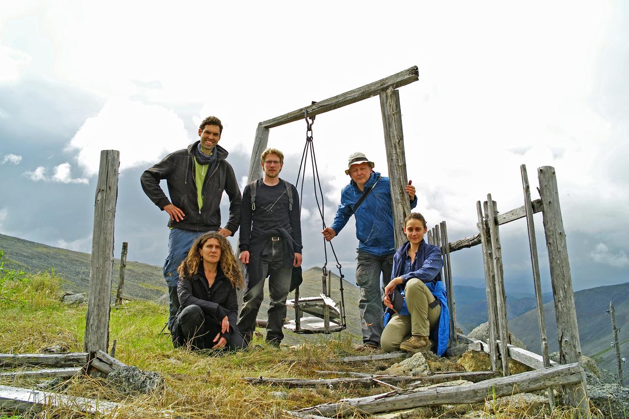 Eine grob gebaute, verwitterte, graubraune leere Kinderschaukel auf einem hoch in den Bergen liegenden, verlassenen Gelände. Rundherum kaputte Zäune und Holzlatten verstreut auf dem Boden. Im Hintergrund Berge, Täler und darüber ein wolkenverhangener Himmel. Um die Schaukel gruppieren sich fünf Personen.