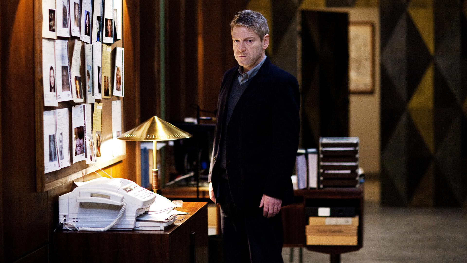 Kommissar Wallander (Kenneth Branagh) steht im Büro vor Fotos der Verdächtigen.