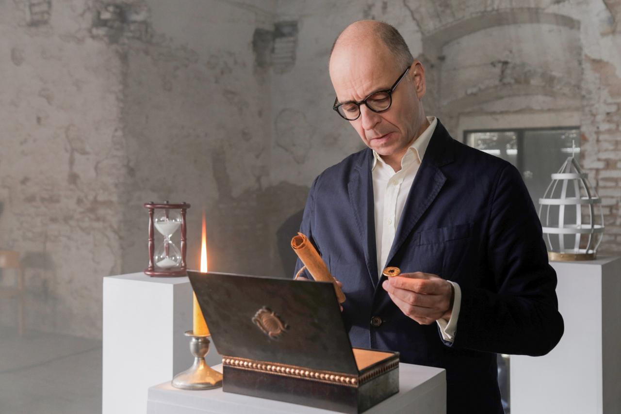 Ein Mann mit Brille betrachtet ein päpstliches Siegel und eine Schriftrolle vor einer geöffneten Truhe. Daneben stehen eine Kerze und eine Sanduhr in einem spärlich beleuchteten, historischen Raum.