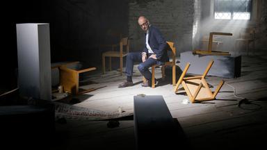 Ein Mann in Anzug sitzt nachdenklich auf einem Stuhl in einem dunklen Raum mit umgestürzten Möbeln und diffusem Licht. Eine Lichtquelle fällt durch ein vergittertes Fenster auf ihn.