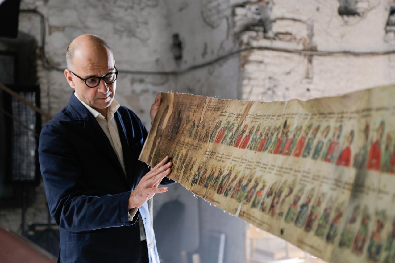 Ein Mann mit Brille hält in einem historischen Raum eine lange, illustrierte Schriftrolle mit Porträts und Text. Er betrachtet sie konzentriert im Lichtschein.