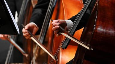 Nahaufnahme der Hände von drei Männern, die in einem Orchester hintereinander stehen und einen Bogen über ihren Kontrabass streichen.