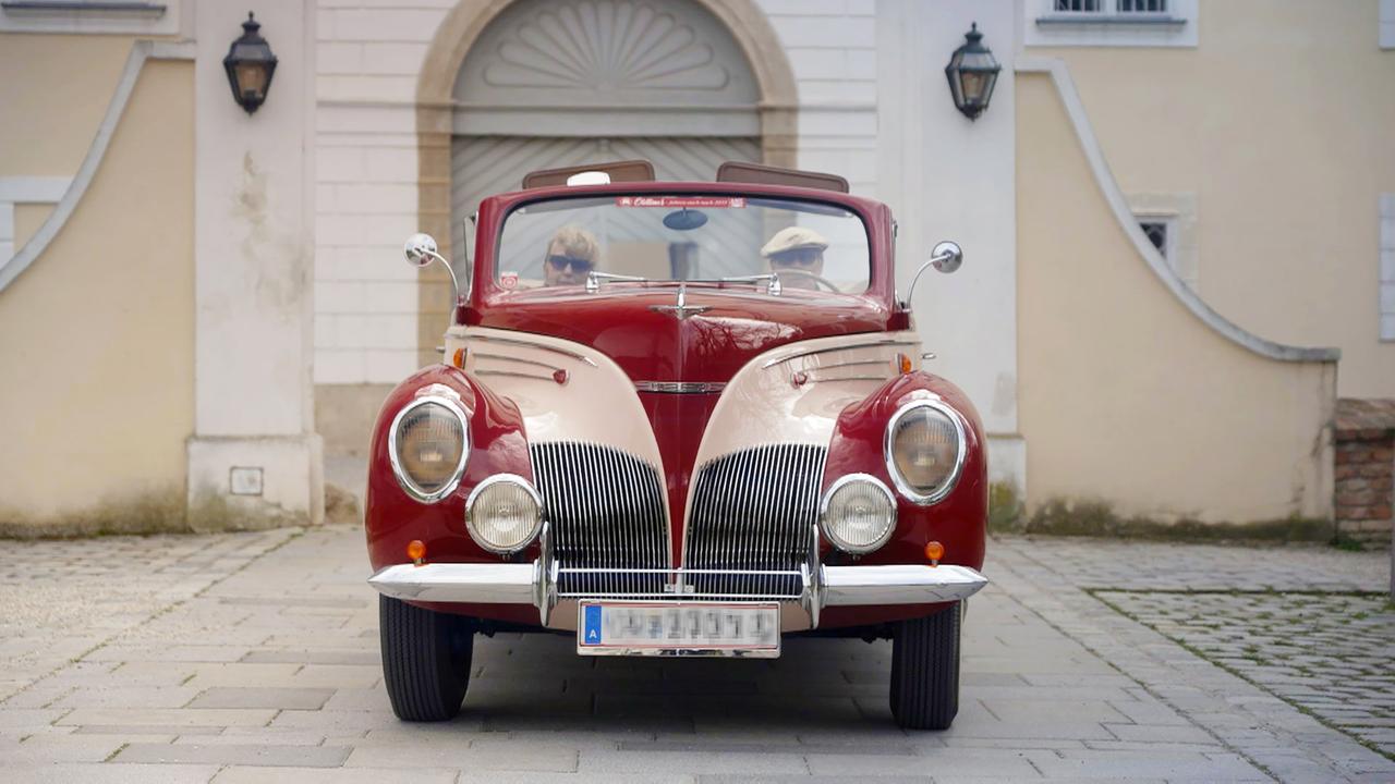 Das Bild zeigt ein rotes und sandfarbenes Oldtimerfahrzeug, einen Lincoln Zephyr aus dem Baujahr 1939, das frontal betrachtet wird. Das Auto hat eine markante Kühlergrillstruktur mit vielen vertikalen Lamellen. Die Scheinwerfer sind rund und weisen ein klassisches Design auf.   Im Fahrer- und Beifahrersitz sitzen zwei Personen, die Sonnebrillen tragen. Die Frau auf der linken Seite hat lockige Haare. Der Mann auf der rechten Seite trägt einen hellen Hut. Im Hintergrund ist eine historische Gebäudefassade zu sehen, die zu einem Schloss oder einem ähnlich alten Bauwerk gehören könnte. Die Fassade ist hell gestrichen und es befinden sich zwei Laternen an der Wand. Der Boden besteht aus Steinen, die ordentlich verlegt sind.   Das Bild vermittelt ein Gefühl von Nostalgie und zeigt die Leidenschaft für Oldtimer und Sammlerstücke.