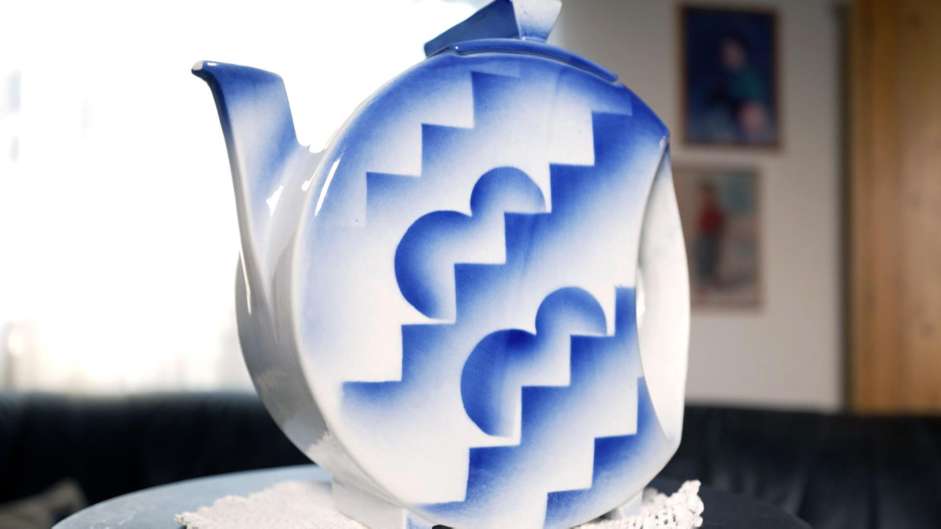 Das Bild zeigt eine dekorative Kanne, die auf einem runden Tisch steht. Die Kanne ist aus Keramik und hat eine weiße Grundfarbe, die mit blauen Mustern verziert ist. Die Muster bestehen aus geometrischen Formen, die an Zickzacklinien und Mondformen erinnern. Die Kanne hat einen länglichen Ausguss und einen kleinen Deckel.   Im Hintergrund ist eine unscharfe, helle Wand zu sehen, die wahrscheinlich ein Fenster darstellt, da Licht hereinströmt. Auf der linken Seite des Bildes ist eine schwarze Couch sichtbar, die mit einem weißen, filigranen Tuch gedeckt ist. An der Wand im Hintergrund hängen einige Rahmen mit Bildern, die jedoch nicht gut zu erkennen sind.   Das Bild vermittelt einen Eindruck von ebenso ästhetischen als auch kurvenreichen Linien und lädt zur Entdeckung der Sammlung ein.
