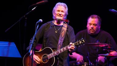 Kris Kristofferson mit Gitarre auf der Bühne. Hinter ihm ist ein Schlagzeuger zu sehen.