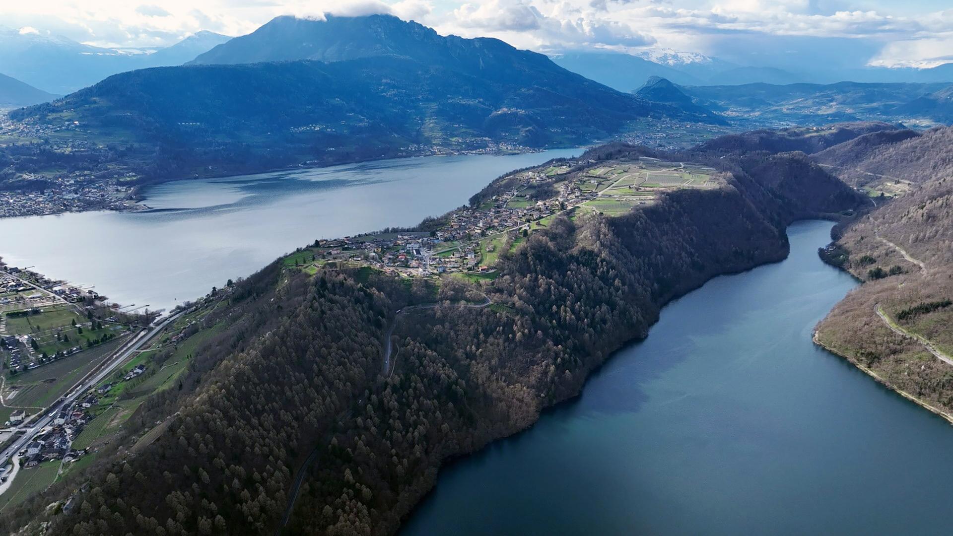 Das Bild zeigt eine Luftaufnahme von zwei Seen, dem Levicosee und dem Caldonazzosee, umgeben von Hügeln und Bergen. Im Vordergrund sind bewaldete Flächen und Siedlungen zu sehen, während im Hintergrund die Berglandschaft durch Wolken partially verdeckt wird.