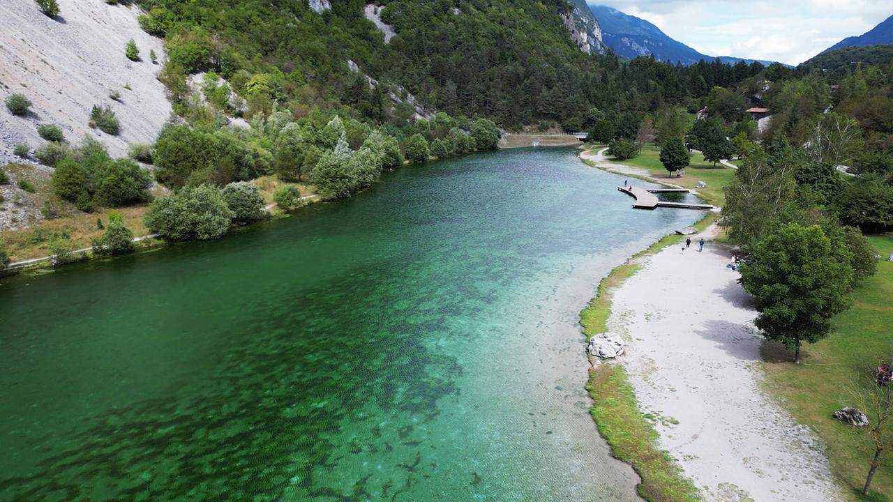 Das Bild zeigt den Lago di Nembia im Trentino. Das Wasser hat eine smaragdgrüne Farbe, umgeben von Bäumen und einer Sandfläche. Am Ufer sind Spaziergänger und eine Holzplattform zu sehen.