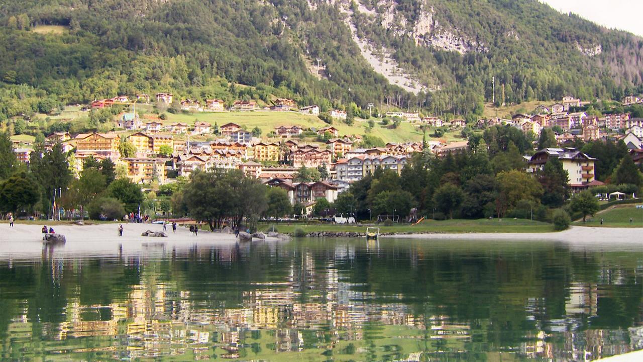 Das Bild zeigt den Molvenosee im Trentino, umgeben von Bergen. Im Vordergrund ist der ruhige See mit einer sandigen Uferzone und einigen Menschen zu sehen. Im Hintergrund sind bunte Gebäude und Wälder.