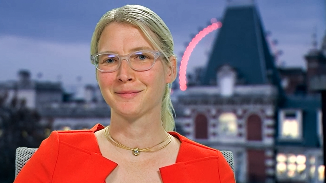 Eine Frau mit zu einem Zopf zurückgebundenen langen Haaren, mit Brille und Goldkette