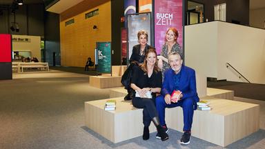 Moderator Gert Scobel und Sandra Kegel sitzen auf einem Podest. Barbara Vinken und Katrin Schumacher sitzen etwas erhöht hinter Scobel und Kegel ebenenfalls auf einem Podest. Im Hintergrund sind Banner der Buchzeit, des Literarischen Quartetts und der Kulturzeit im Foyer der Frankfurter Buchmesse zu sehen. 