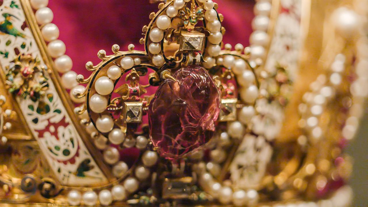 Das Bild zeigt ein Detail der Kaiserkrone, die Teil der Kaiserlichen Schatzkammer in der Wiener Hofburg ist. Die Krone ist aufwändig verziert mit goldenen und silbernen Elementen sowie einer Vielzahl von Perlen. Im Zentrum befindet sich ein großer, rosafarbener Edelstein. Es sind auch kleine, quadratische Steine um den Edelstein herum sichtbar, die einen glitzernden Effekt erzeugen. Der Hintergrund der Krone ist mit einem tiefen Rotton ausgekleidet, der einen Kontrast zu den goldenen Verzierungen bildet. Die Bildkomposition hebt die filigrane Handwerkskunst und den Reichtum der Kronjuwelen hervor.