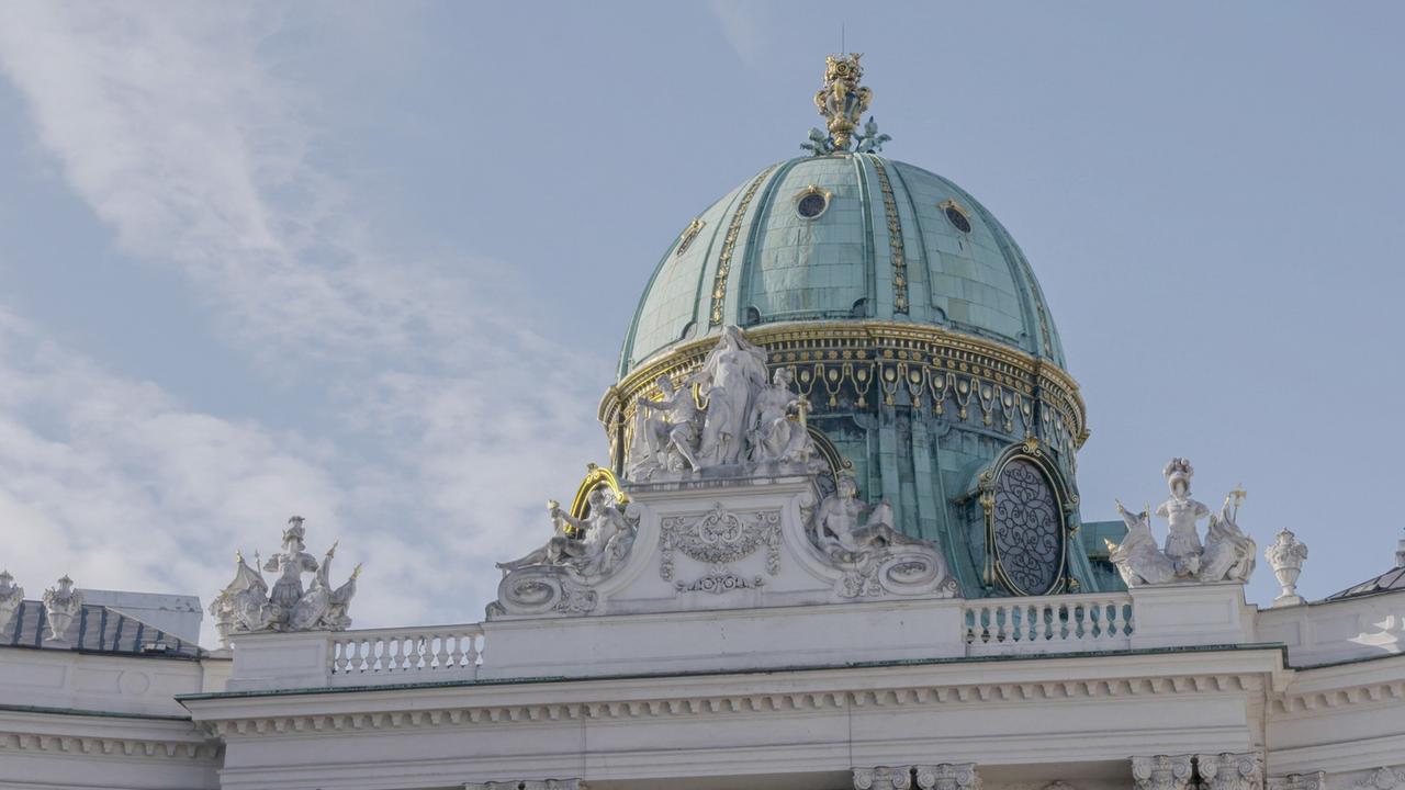Das Bild zeigt die Kuppel des Michaelertors der Wiener Hofburg. Die Kuppel ist aus grünem Kupfer und weist goldene Verzierungen auf. An der Spitze der Kuppel befindet sich eine weitere Skulptur, umgeben von mehreren anderen dekorativen Elementen. Im Hintergrund sind helle Wolken und ein blauer Himmel sichtbar. Das Gebäude ist in einem klassischen architektonischen Stil gehalten, mit geschwungenen Linien und reichhaltigen Details an den Fassaden.