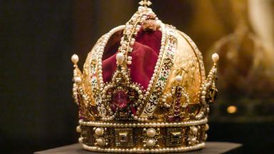 Das Bild zeigt eine prächtige Krone, die als Kaiserkrone bekannt ist. Sie ist aus goldfarbenem Material gefertigt und reich verziert mit Perlen, Edelsteinen und kunstvollen Verzierungen. In der Mitte der Krone befindet sich ein großer, rosa Edelstein, umgeben von weiteren glitzernden Steinen und schimmernden Perlen. Der obere Teil der Krone weist eine geschwungene Form auf und ist mit einer feinen Goldverzierung sowie zahlreichen kleinen Schmucksteinen dekoriert. Der Hintergrund ist dunkel und unscharf, wodurch die Krone im Vordergrund stark hervorsticht.   Diese Krone gehört zur Kaiserlichen Schatzkammer in der Wiener Hofburg und hat eine historische Bedeutung im Zusammenhang mit der Habsburgermonarchie.