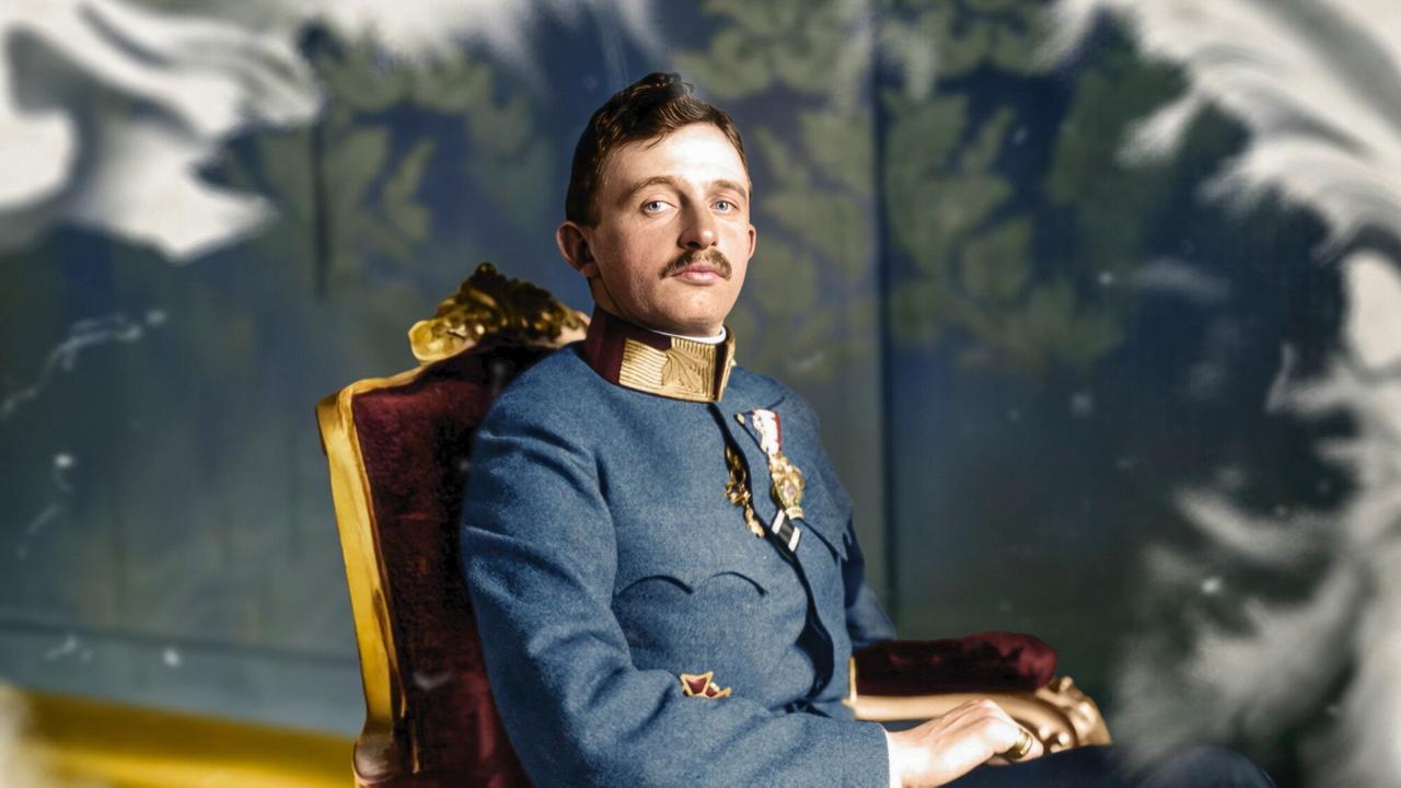 Das Bild zeigt einen jungen Mann in einer formellen Uniform. Er sitzt auf einem roten, gepolsterten Sessel mit goldenen Akzenten. Der Mann hat einen schmalen Oberkörper und trägt eine elegante Militäruniform in Blau, die mit verschiedenen Abzeichen und Orden geschmückt ist. Er hat kurz geschnittenes, braunes Haar und einen kleinen, gepflegten Schnurrbart.   Der Hintergrund des Bildes ist unscharf, mit einer weichen, lichteinfallenden Textur, die die Aufmerksamkeit auf den Mann lenkt. Er schaut direkt in die Kamera und hat einen ernsten, nachdenklichen Gesichtsausdruck. Die Lichtverhältnisse sind warm und betonen die Farben der Uniform sowie die Struktur des Sessels.  Das Bild ist Teil des historischen Kontextes des Films „Der Raub der Kronjuwelen“, der sich mit dem Verschwinden der Kronjuwelen der Habsburgermonarchie im Jahr 1918 beschäftigt.