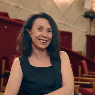 Shermin Langhoff sitzt in einem Theater auf einem Stuhl aus roten Samt. Sie lächelt in die Kamara.