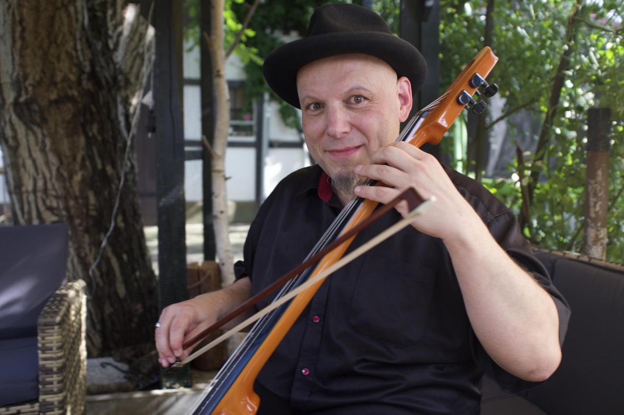 Mann in schwarzem Hemd und mit Hut hält Streichinstrument in der Hand.