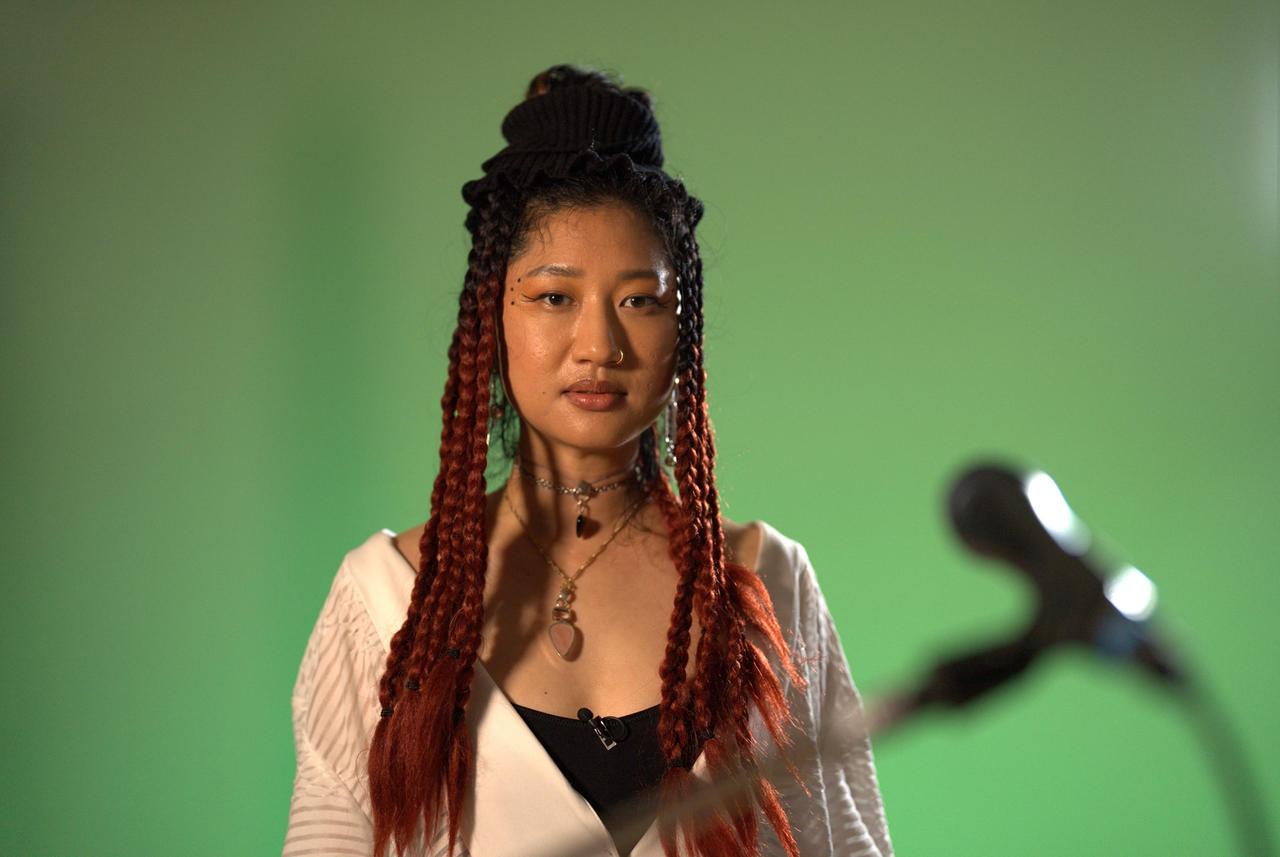 Frau mit langen geflochtenen Zöpfen vor grünem Hintergrund. Im Vordergrund unscharf ein Mikro zu erkennen.