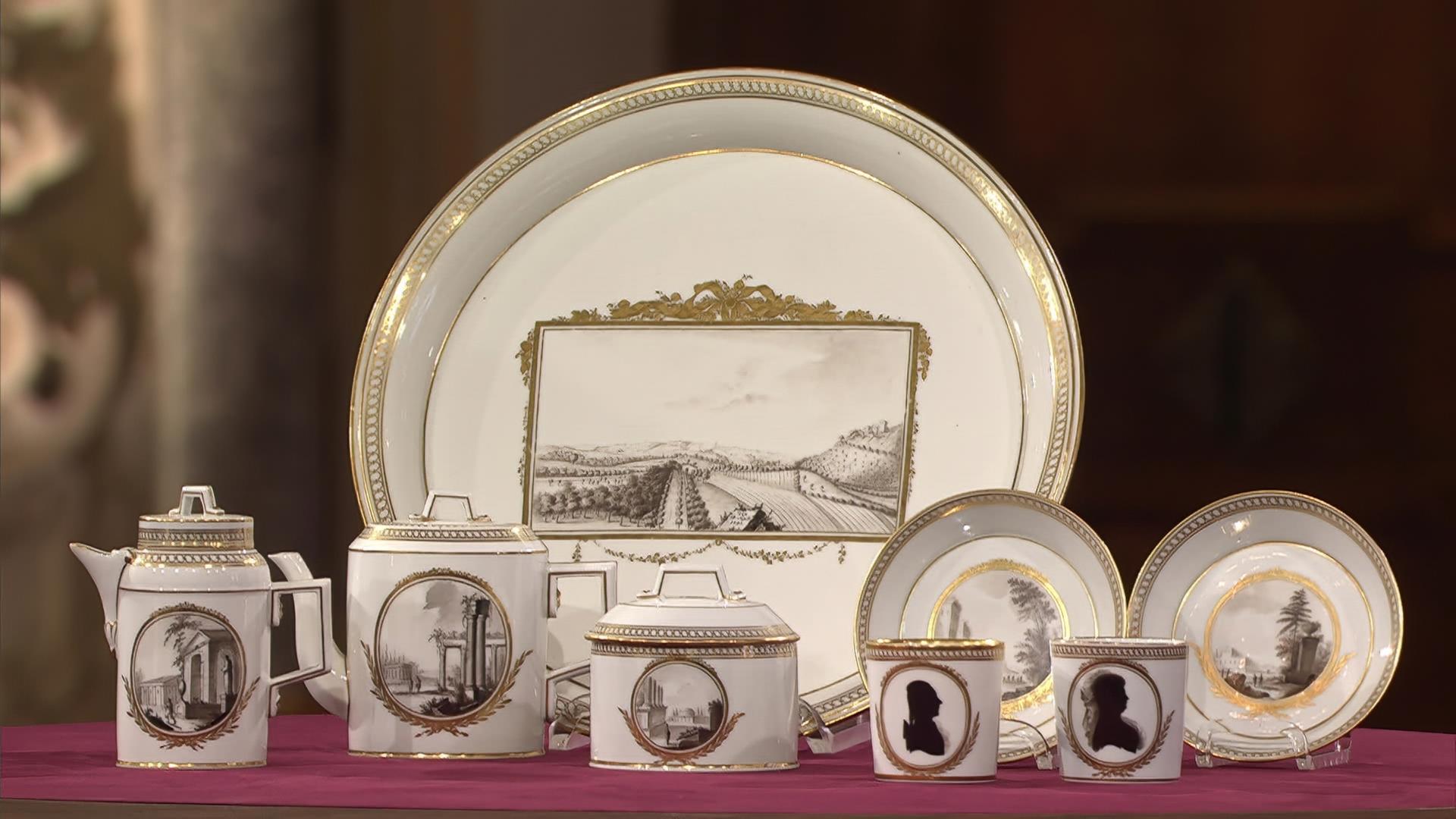 Ein weiß-goldenes Porzellan-Geschirr, auf das schwarze Landschaftsmotive und Büsten gedruckt sind.