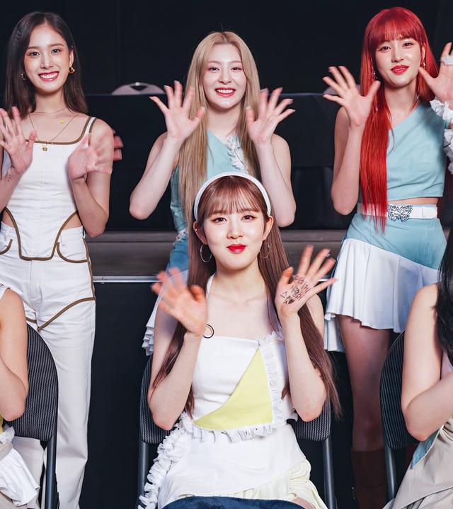 Sechs asiatische, junge Frauen. Drei vorne sitzend, drei dahinter stehend. Sie lächeln in die Kamera und winken mit beiden Händen. Sie haben alle lange glatte Haare, drei davon schwarze, eine braune, eine blonde und eine rote. Sie tragen roten Lippenstift. Die Kleidung ist vornehmlich weiß mit teilweise gelben, türkisen und hellbraunen Akzenten. 