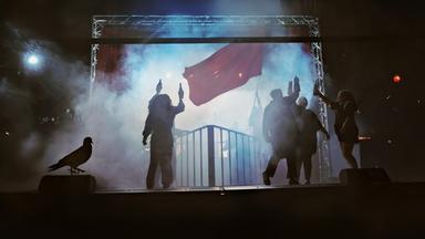 Theaterinszenierung mit roter Fahne und rauchgeschwängerter Atmosphäre, mehrere Menschen mit erhobenen Flaschen auf einer Bühne in dunklem Licht