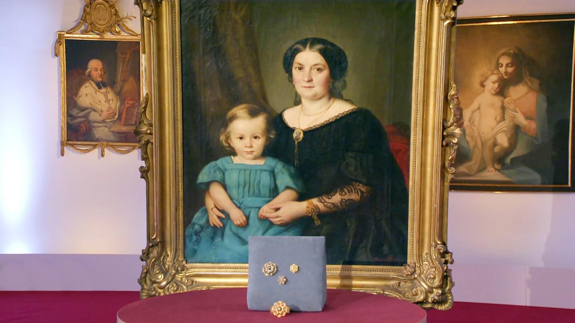 Ein goldgerahmtes Gemälde von einer Frau mit Kind. Davor stehen auf einem Brettchen Orringe, Brosche und ein Ring.