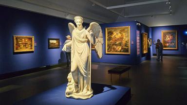 In einem abgedunkelten Ausstellungsraum mit blau gestrichenen Wänden steht in der Mitte eine große Marmorstatue mit Flügeln. An den Wänden hängen Ölgemälde, die ein paar undeutlich erkennbare Besucher betrachten.