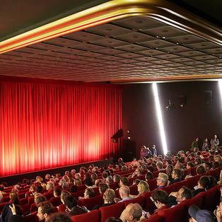 Das Publikum sitzt vor einem roten, geschlossenem Vorhang und wartet auf die Vorstellung. 