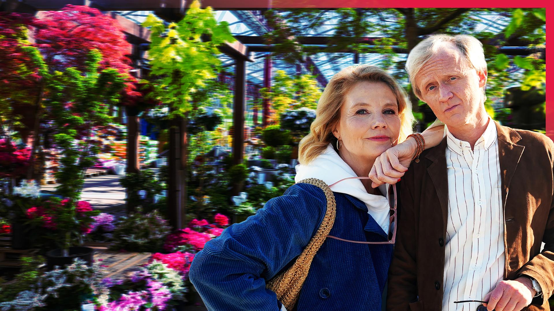 Annette Frier und Pierre M. Krause stehen unter einer Pergola in einer Pflanzenhandlung voller blühender Pflanzen.