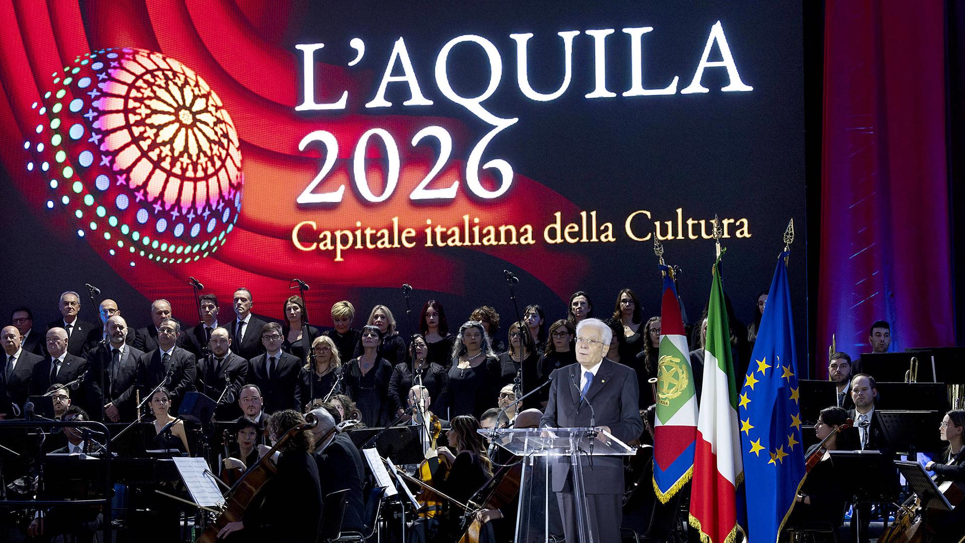 An einem Podest aus Plexiglas neben einem Orchester steht ein Mann und spricht. Im Hintergrund ist groß die Schrift "L'Aquila 2026 Capitale italiana della Cultura auf buntem Hintergrund zu sehen.