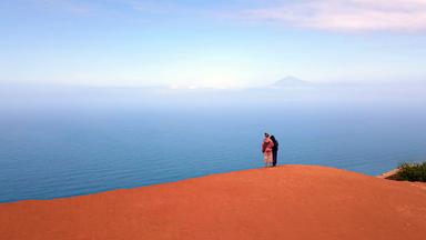 Rückansicht von zwei Menschen, die von Gomera aus in die Ferne blicken. Im Hintergrund ein Berg.