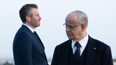 Im Vordergrund steht ein älterer Mann im Anzug mit Brille, der nachdenklich zur Seite schaut. Hinter ihm steht sein Bodyguard, ein Mann mittleren Alters, ebenfalls im Anzug und Knopf im Ohr.