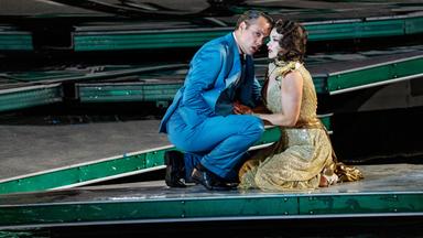 Ein Mann in einem blauen Anzug kniet neben einer sitzenden Frau in einem goldfarbenen Kleid auf der Bühne.