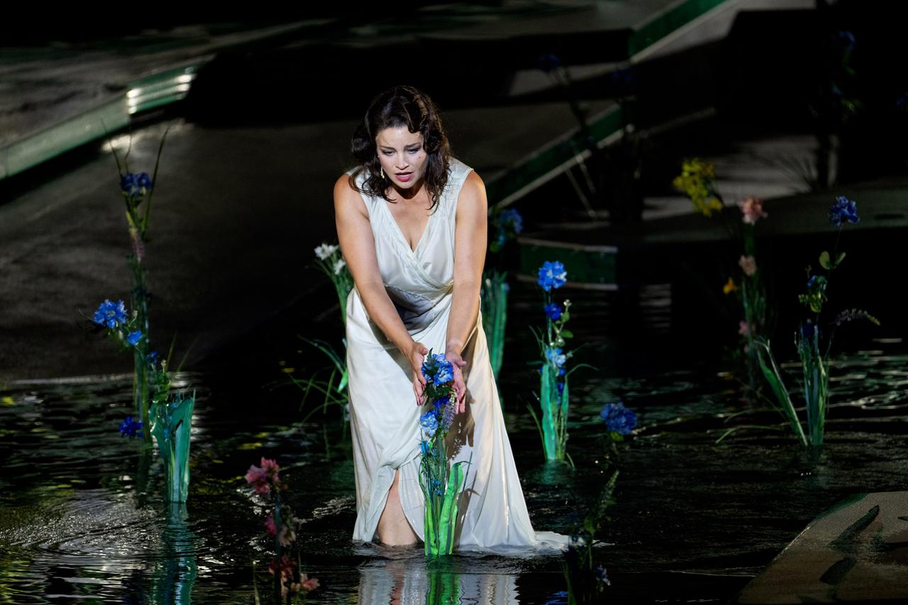 Eine Frau in einem weissen Kleid umfasst blaue Blumen auf der Seebühne, die vor ihr im Wasser wachsen.