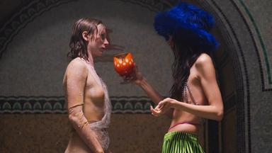 Im Profil sind zwei junge Frauen zu sehen, die sich gegenüber stehen. Eine hat rötliches, nasses Haar und trägt eine Art durchsichtige Schlangenhaut, die andere trägt eine auffällige grüne Hose, ein Kettenoberteil und einen blauen Plüschhut, ihr Haare ist lang und dunkel, ihr Gesicht sieht man nicht. Sie hält der änderen einen orangefarbenen Kelch hin.