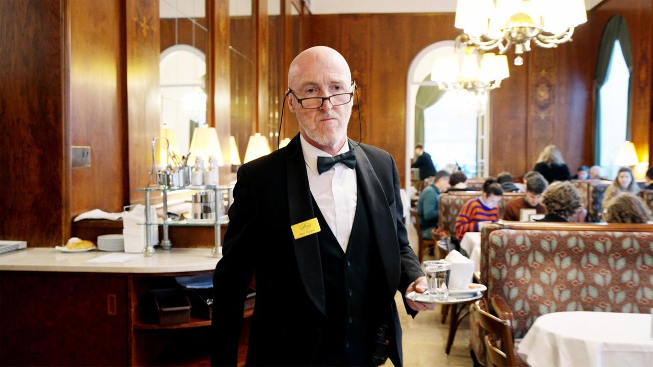 Das Bild zeigt einen Oberkellner in eleganter Kleidung, der ein Serviertablett mit einem Glas Wasser und einer Tasse Kaffee in der Hand hält. Er trägt einen klassischen schwarzen Anzug mit einer weißen Shirt und einer schwarzen Fliege. Zudem hat er eine Namensschilder auf seiner Jacke, auf dem vermutlich der Name "Herr Rudolf" steht.   Der Hintergrund des Bildes zeigt eine stilvolle Café- oder Restaurantumgebung mit warmen Holztönen und mehreren Tischen und Stühlen. An den Tischen sitzen Gäste, einige von ihnen lesen oder unterhalten sich, während andere Essen oder Getränke konsumieren. Große Fenster lassen Tageslicht herein und tragen zur angenehmen Atmosphäre bei. Es sind auch mehrere Lampen zu sehen, die den Raum weiter erhellen.   Die gesamte Szenerie vermittelt ein Gefühl von Eleganz und einem gehobenen Ambiente, das häufig in traditionellen Kaffeehäusern oder Restaurants zu finden ist.