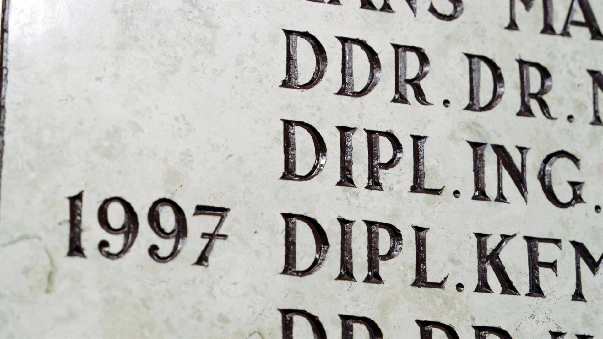 Das Bild zeigt einen Ausschnitt einer Gedenktafel oder Plakette aus einem hellen Material, das möglicherweise in Stein gemeißelt ist. Die Oberfläche der Tafel ist texturiert und zeigt eine Vielzahl von eingravierten Buchstaben und Zahlen.   Die Inschrift enthält akademische Titel, darunter "DDR. DR.", "DIPL. ING." und "DIPL. KFM.", sowie die Jahreszahl "1997". Die Buchstaben sind in Großbuchstaben und zeichnen sich durch eine kräftige, dreidimensionale Form aus, wodurch sie gut lesbar sind. Die Buchstaben scheinen dunkel eingelassen zu sein, was einen starken Kontrast zur hellen Oberfläche schafft.   Die umgebende Fläche der Tafel wirkt rau und hat einige kleine Risse oder Abnutzungen, die auf ein gewisses Alter hindeuten. Das Gesamtbild vermittelt den Eindruck von formeller Ehrung und akademischer Traditionspflege.
