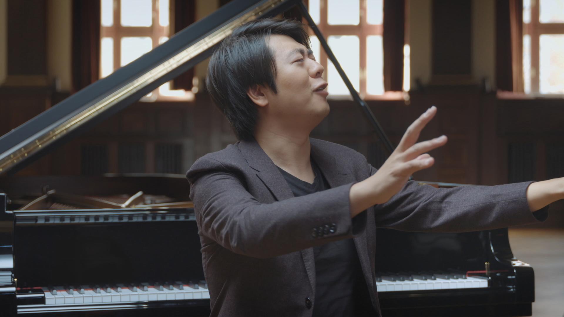 Lang Lang sitzt mit dem Rücken vor einem Flügel. Er hat die Augen geschlossen und den Kopf leicht nach links oben gedreht. Beide Arme sind schulterhoch als ob er dirigieren würde.