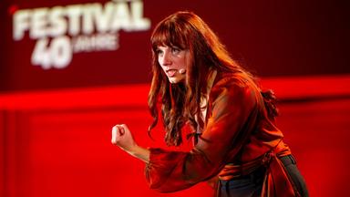Lara Ermer streckt den linken Arm vor sich aus und ballt die Hand zu einer Faust. Sie trägt eine rote Bluse vor rot beleuchtetem Hintergrund. Sie schaut zum linken Bildrand.